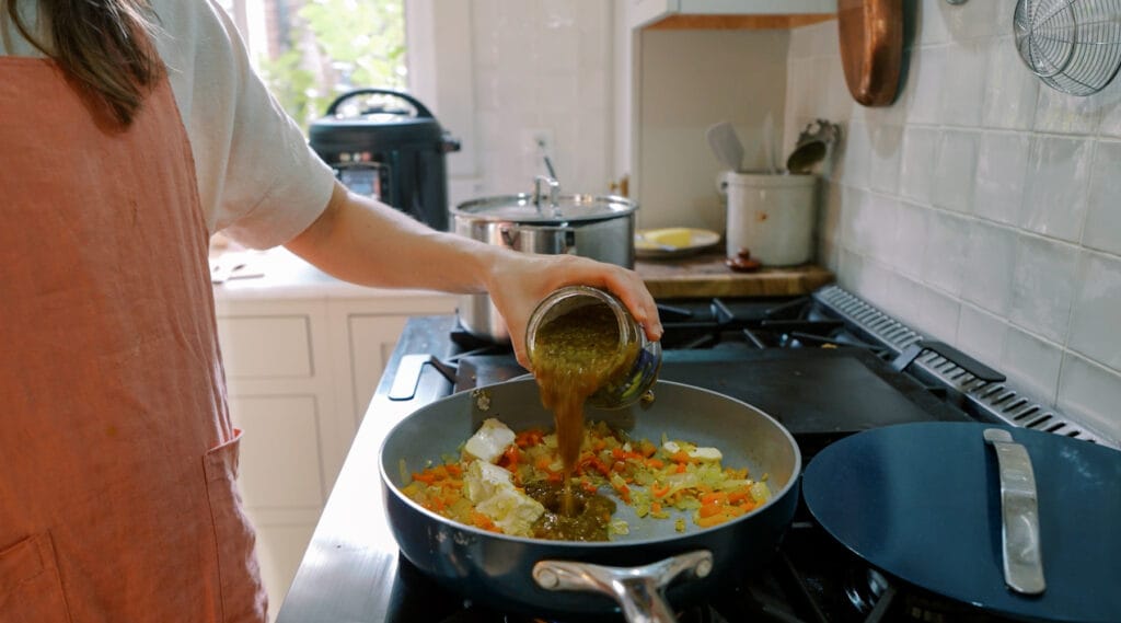 Person pouring salsa verde into a skillet with sautéed onions, peppers, and cream cheese.