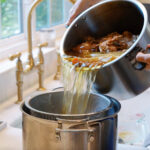 Roasted chicken bones, onions, celery, and garlic being added to an Instant Pot for homemade bone broth.