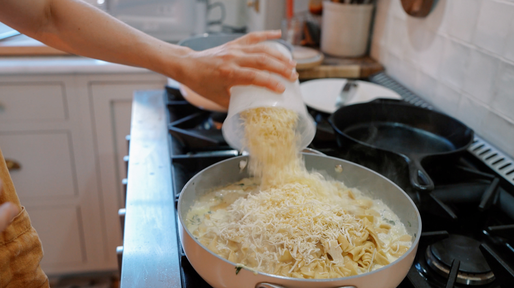 pouring parmesan cheese over pasta alfredo in a skillet
